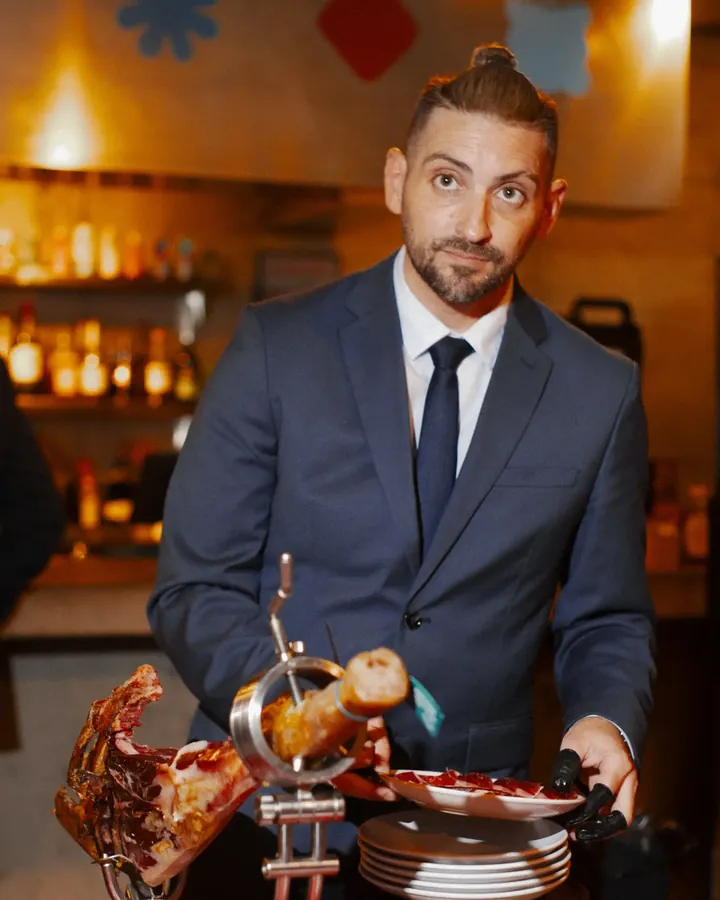Host in a navy suit carving jamón from the leg at the bar, plates stacked beside him