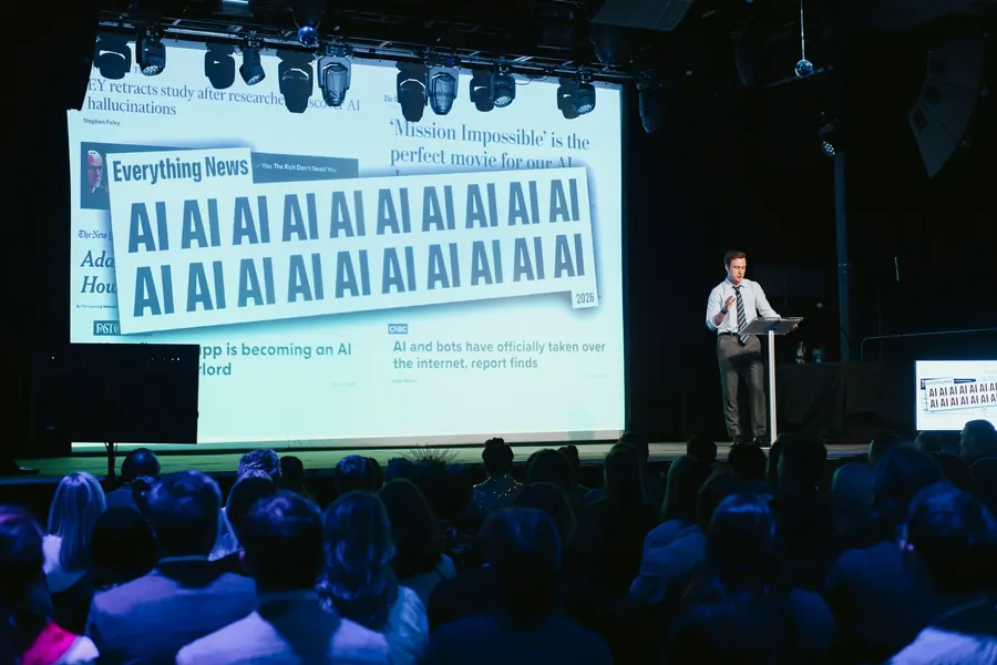 Speaker on stage in front of a wall-sized screen of AI headlines, room packed to the back